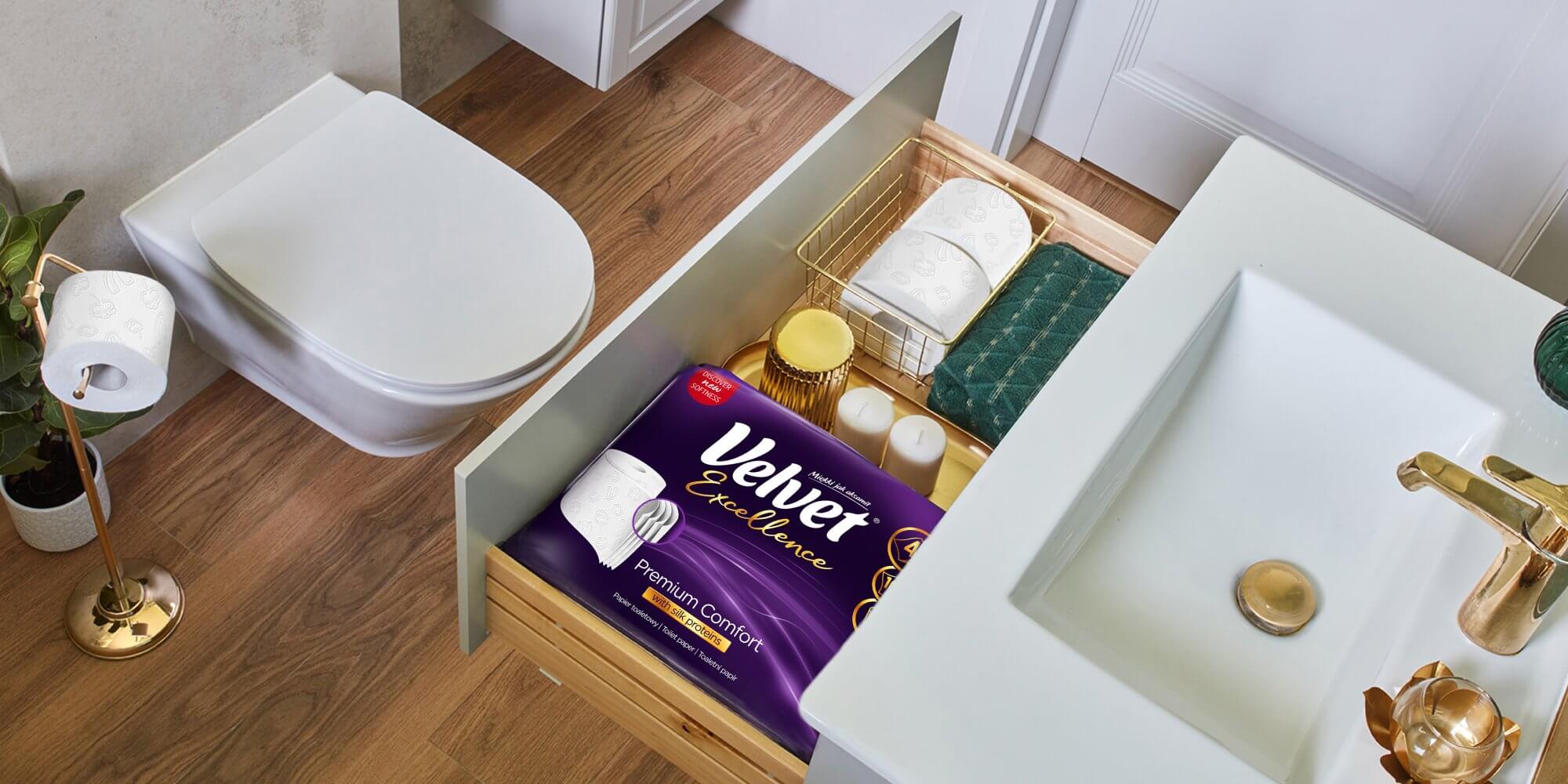Nowoczesna łazienka z otwartą szufladą, w której znajdują się produkty Velvet Excellence – papier toaletowy Premium Comfort w fioletowym opakowaniu, dodatkowe rolki w koszyku oraz akcesoria łazienkowe.